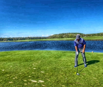Golfer preparing to tee off on a lush green course by a serene lake, illustrating the outdoor golfing experience emphasized in indoor golf simulator discussions.