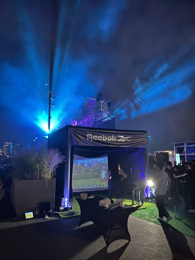 Reebok-branded golf simulator setup at night event with vibrant blue and purple lights, featuring a projection of a golf course scene, surrounded by engaged attendees and a table with golf accessories.
