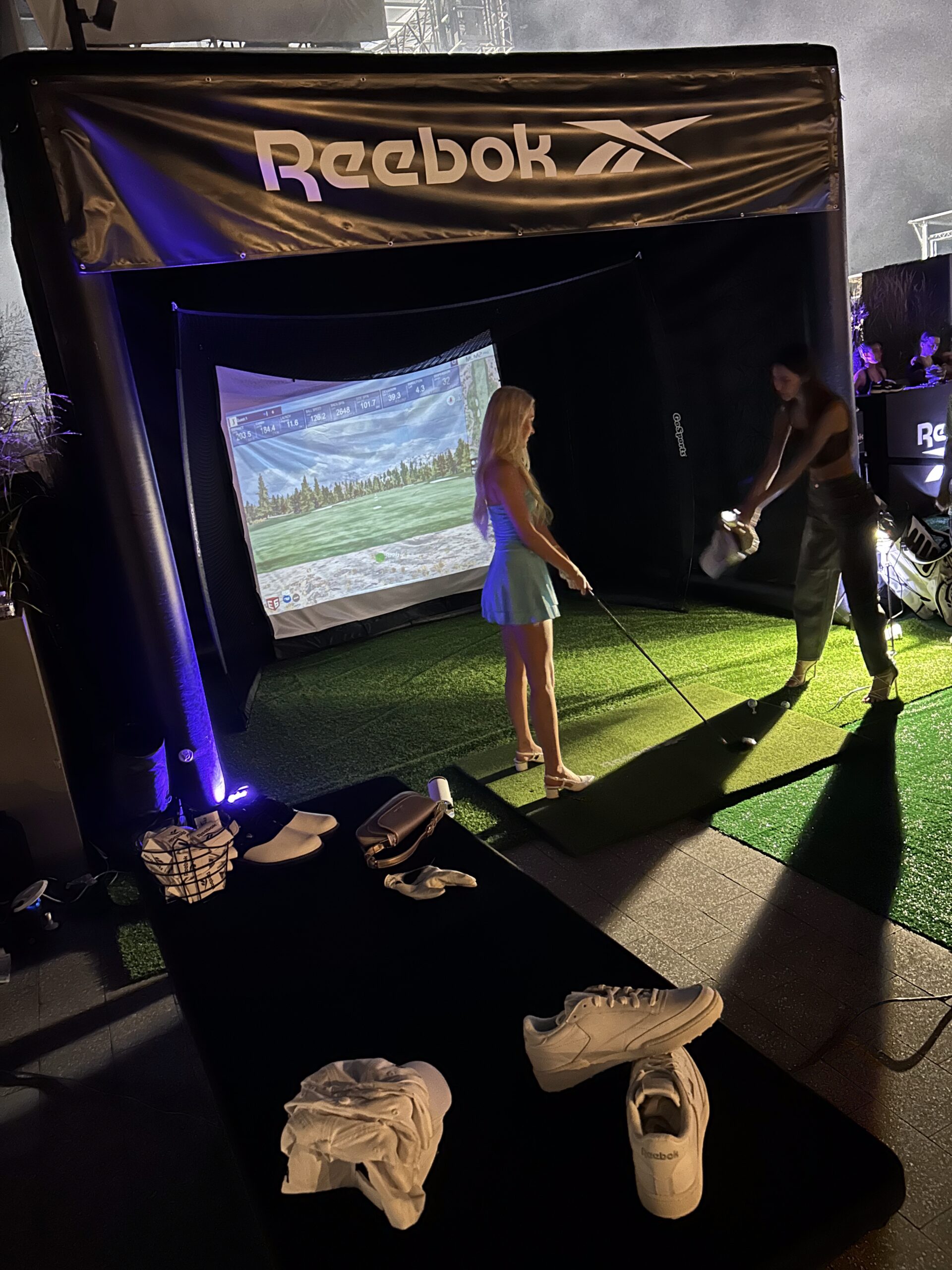 Indoor golf simulator with Reebok branding, two women engaging