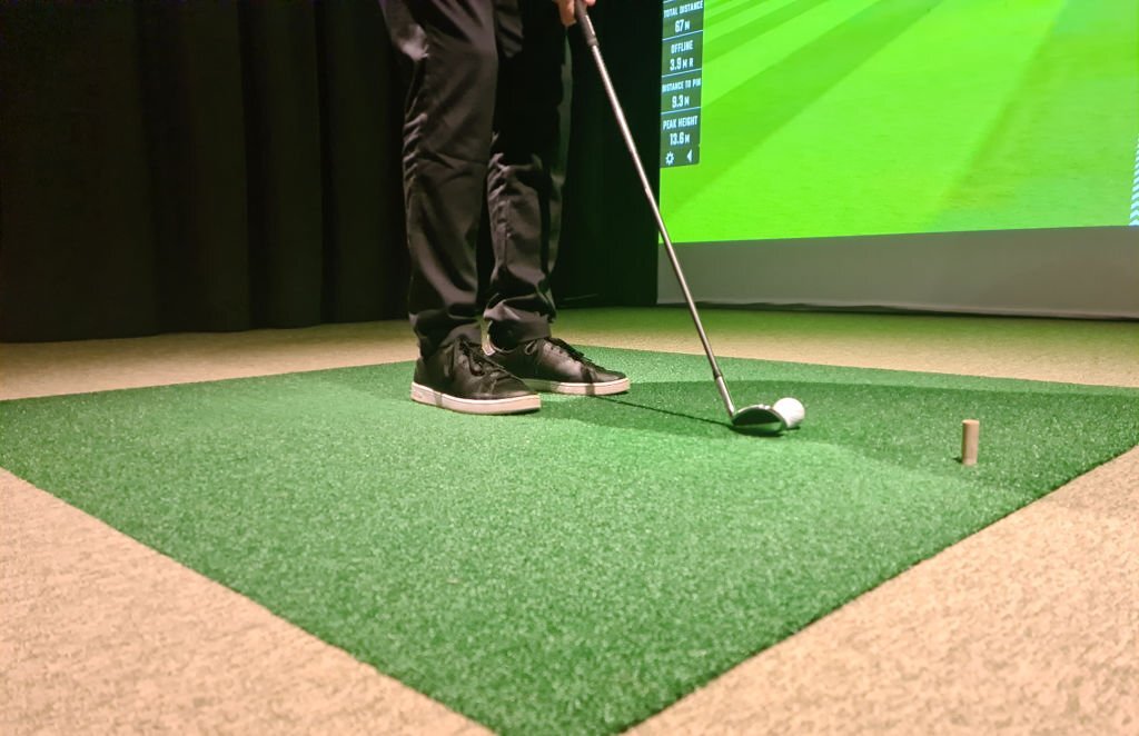 Golfer preparing to swing a club on a synthetic grass mat in an indoor golf simulator, with a digital screen displaying performance metrics in the background.