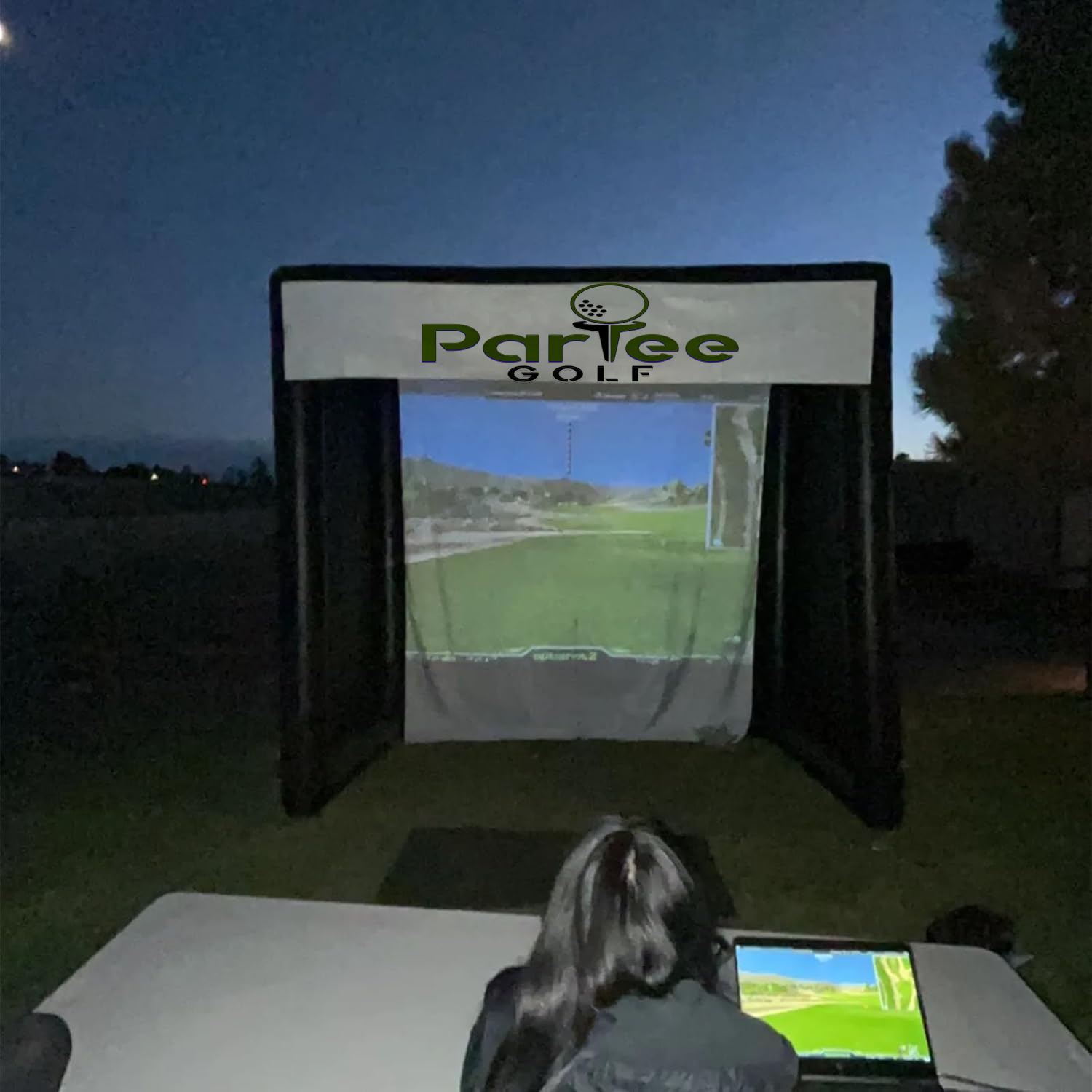 Portable golf simulator setup by ParTee Golf, featuring a projection screen displaying a golf course, with a user engaging at a table in an outdoor evening setting.