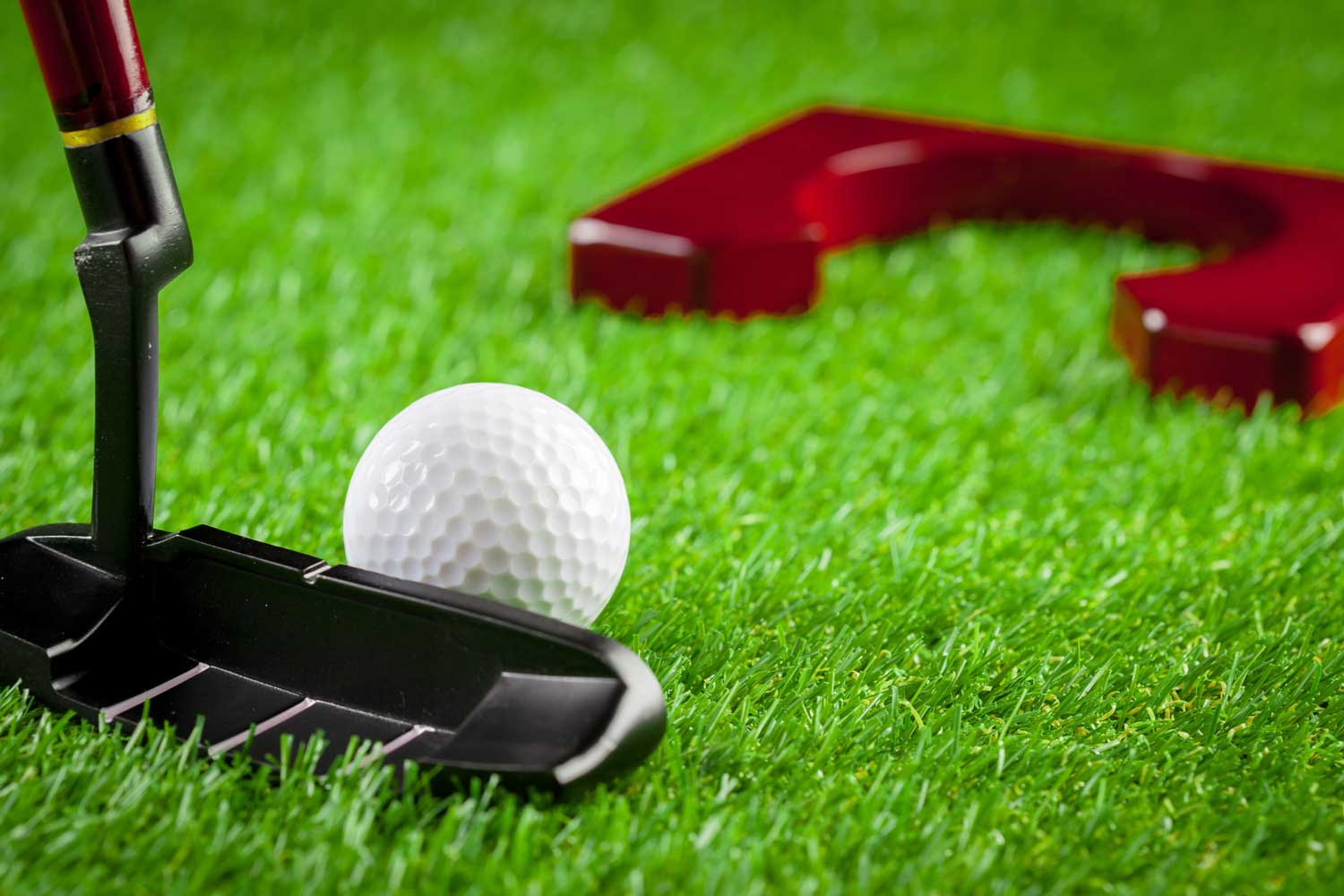 Black golf putter and white golf ball on green artificial grass, with a red mini-golf obstacle in the background, illustrating indoor golf equipment for practice and skill improvement.